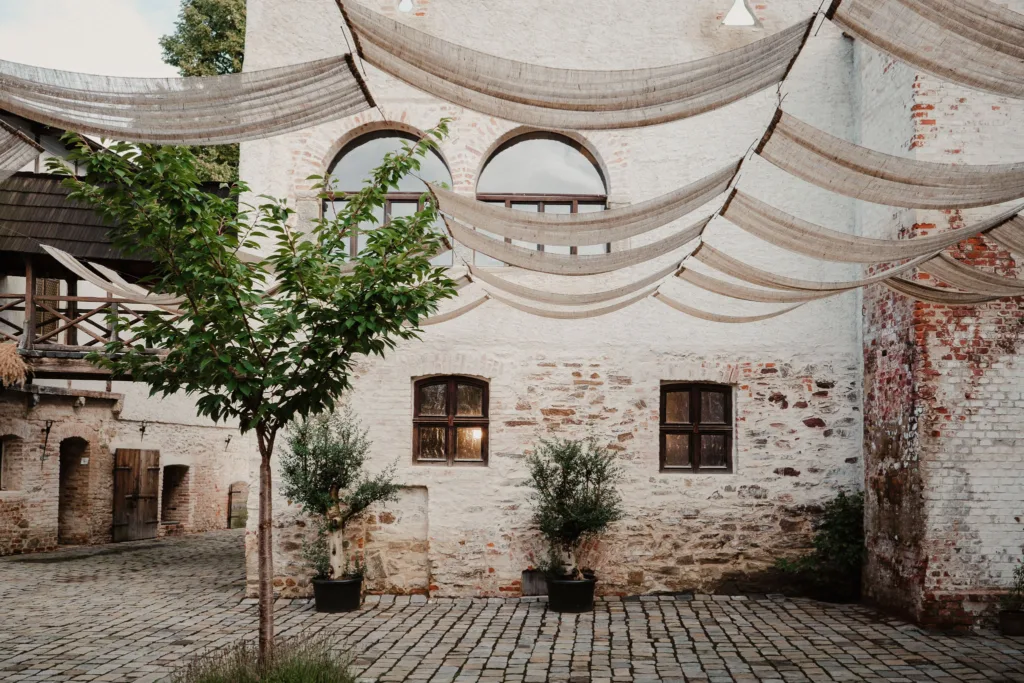 Venkovní nádvoří Za Vratama se stínícími plachtami a olivovníky, které vytváří toskánskou atmosféru.